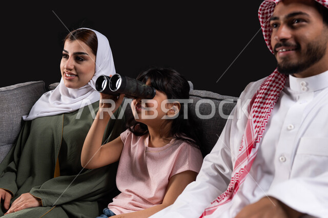 دعم مواهب الأطفال وتنميه قدراتهم على حب الإستكشاف والتطلع للمستقبل، بورتريه مقرب لعائله عربيه خليجيه سعوديه تجلس على الأريكه، طفله سعوديه تحمل المنظار وتقوم بالنظر إلى شيء ما، الاستمتاع والسرور بالجلسات العائليه الهادئه، خلفيه سوداء.