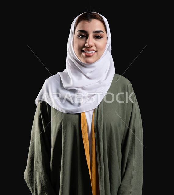 القيام بجلسه تصوير من أجل إلتقاط صور شخصيه لوثائق رسميه، الأهتمام بالجمال والأناقه والمظهر الخارجي، بورتريه مقرب لسيده عربيه خليجيه سعوديه محجبه مبتسمه ترتدي عباءه خضراء مطرزه تنظر إلى الكاميرا، ايماءات وجه تدل على الأنوثه والجمال، خلفيه سوداء.