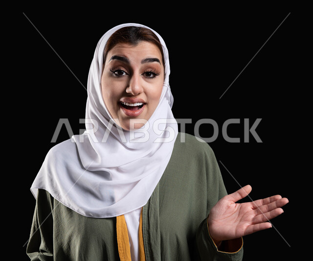 عدم المعرفه والجهل بالأسباب، ايماءات المفاجئه والدهشه والاستغراب، بورتريه مقرب لإمرأه عربيه خليجيه سعوديه محجبه ترتدي عباءه خضراء مطرزه تبتسم وتقوم بالنظر إلى الكاميرا، الإستفهام والسؤال عن شيء ما، خلفيه سوداء.