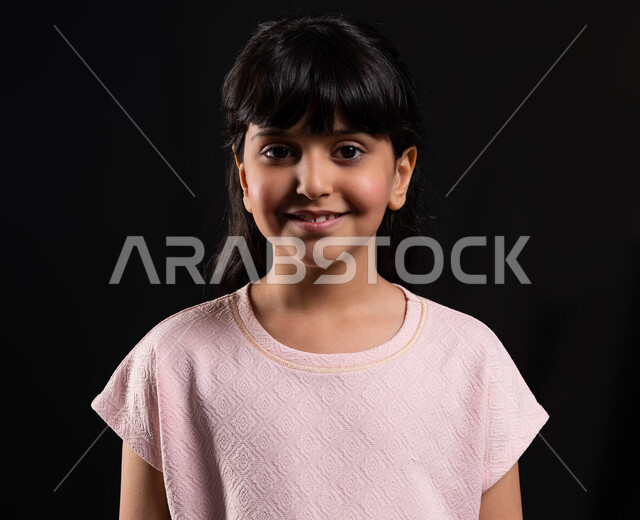 ايماءات وتعابير وجه تدل على السعاده والسرور، الوقوف أمام الكاميرا والنظر بثقه إستعدادا لإلتقاط صوره شخصيه للوثائق الرسميه، بورتريه مقرب لفتاة عربيه خليجيه سعوديه مبتسمه ترتدي الزي الكاجوال، خلفيه سوداء.