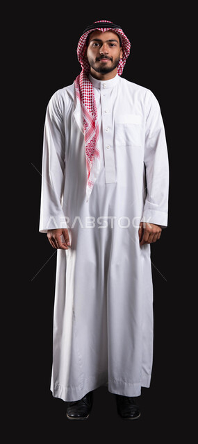 الإهتمام بالمظهر الخارجي، بورتريه لرجل عربي خليجي سعودي يرتدي الشماغ والثوب التقليدي يقوم بالنظر إلى الكاميرا، ايماءات وجه تدل على الثقه بالنفس، صوره الجسم بالطول الكامل، خلفيه سوداء.