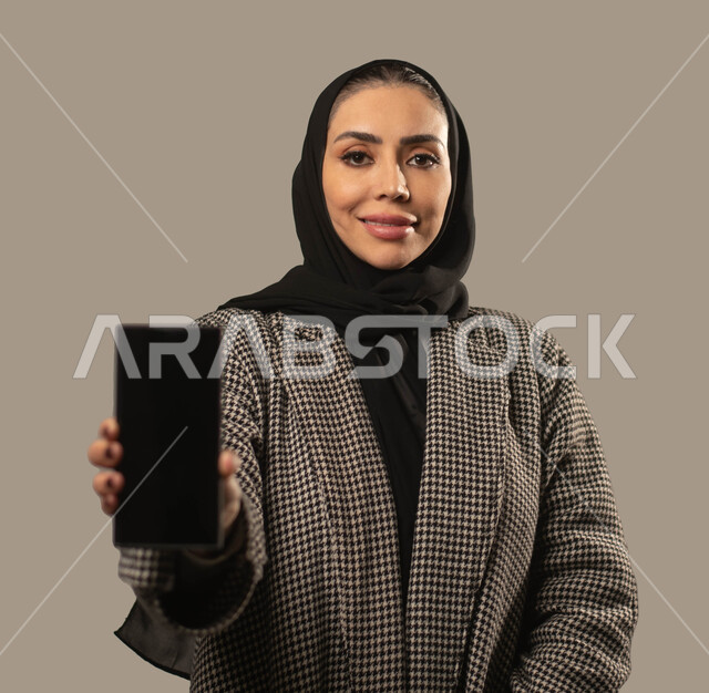 Presentation of mobile applications, e-marketing using technology and technology, a close-up portrait of a smiling Saudi Arabian Gulf woman wearing a hijab, holding a mobile phone with a blank black screen, looking at the camera, a gray background