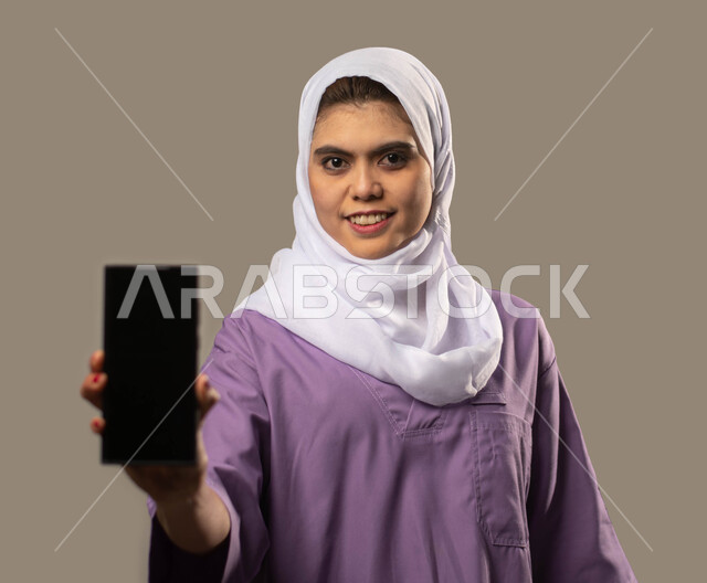 الخدمات الإلكترونيه المتقدمه لدعم و توفير الخادمات والعاملات المرخصة والمؤهله، منصات إلكترونية لاستقدام العماله المنزليه المدربة، بورتريه مقرب لعاملة نظافة افريقية مبتسمة ترتدي الحجاب تحمل هاتف بشاشة سوداء فارغه، مهن ووظائف نسائية، خلفية رمادية