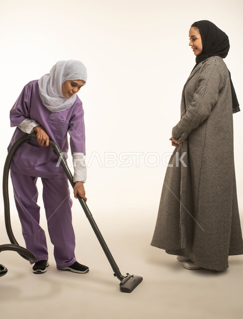 ايماءات السعادة والرضا عن جودة خدمة العاملة، توفير خدمات العناية بالمنزل والنظافة والترتيب، بورتريه لسيدة عربية خليجية سعودية تعطي الارشادات والتوجيهات لخادمة افريقية تقوم باستخدام المكنسة الكهربائية، استقدام العمالة الخارجية المرخصة والمؤهلة، خلفية بيضاء