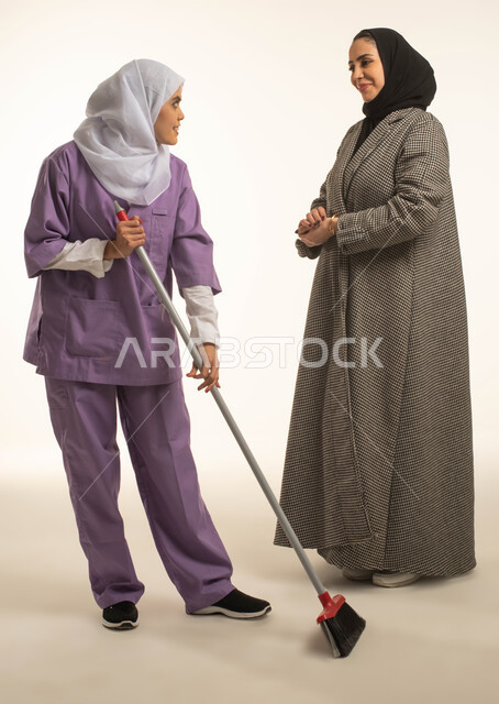 تأمين خدمات العناية بالمنزل والنظافة والترتيب، اصغاء العاملة المنزليه الى طلبات ربة المنزل، بورتريه لسيدة عربية خليجية سعودية تعطي الارشادات والتوجيهات لخادمة افريقية تقوم باستخدام المكنسة اليدوية، استقدام العمالة الخارجية المرخصة والمؤهلة، خلفية بيضاء