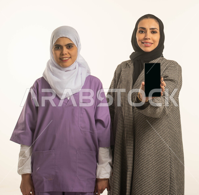 منصات إلكترونية لاستقدام العماله المنزليه المدربة، الخدمات الإلكترونيه المتقدمه لدعم وتوفير الخادمات والعاملات المرخصة والمؤهله، بورتريه لسيدة محجبة تحمل بيدها هاتف بشاشة سوداء فارغة وعاملة نظافة افريقية مبتسمة ترتدي الحجاب، مهن ووظائف نسائية، خلفية بيضاء