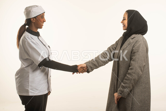 تشغيل الطباخات المرخصة والمؤهلة من الشركات والمكاتب السعوديه، بورتريه مقرب لسيدة عربية خليجية سعودية محجبة تقوم بمصافحة طباخة افريقية ترتدي زي الشيف، الاستقبال والترحيب بالعاملات المنزليه المدربة، أعمال ومهن ووظائف نسائية، خلفية بيضاء