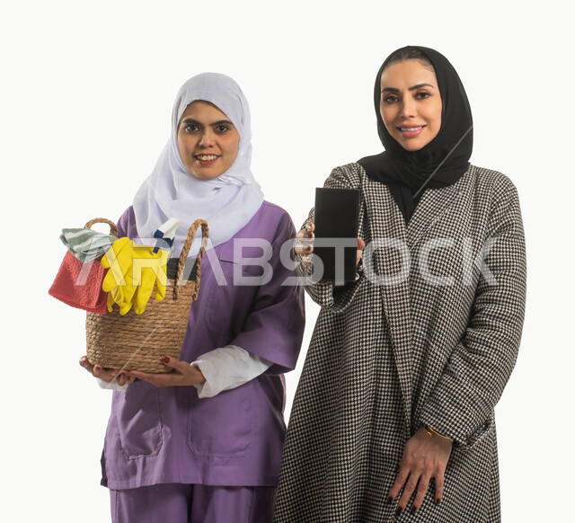 منصات إلكترونية لاستقدام العماله المنزليه المدربة، الخدمات الإلكترونيه المتقدمه لدعم وتوفير الخادمات المرخصة والمؤهله، بورتريه مقرب لسيدة محجبة تحمل بيدها هاتف بشاشة سوداء فارغة وعاملة نظافة افريقية مبتسمة ترتدي الحجاب، مهن ووظائف نسائية، خلفية بيضاء