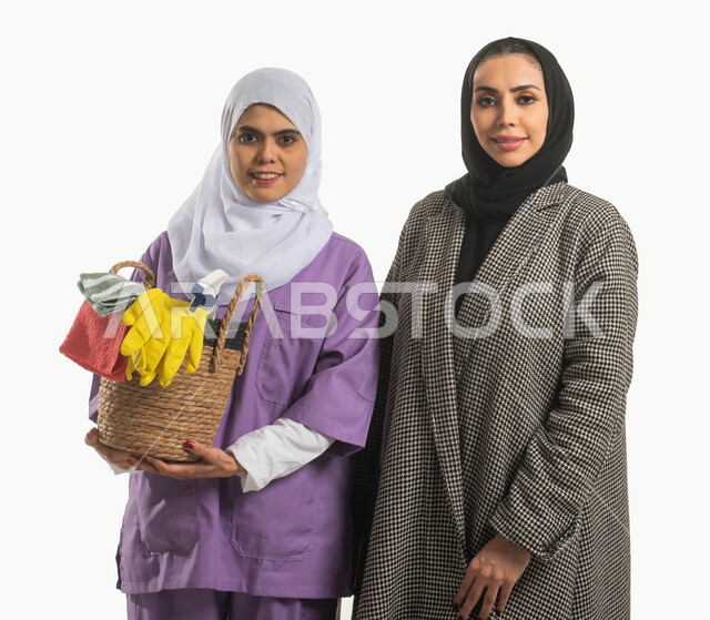 استقدام العماله المنزليه المدربة وتوفير الخادمات والعاملات المرخصة والمؤهله، بورتريه مقرب لسيدة مبتسمة محجبة تنظر إلى الكاميرا وعاملة نظافة افريقية ترتدي الحجاب تمسك بيدها سلة قش بأدوات تنظيف المنزل، مهن ووظائف نسائية، خلفية بيضاء