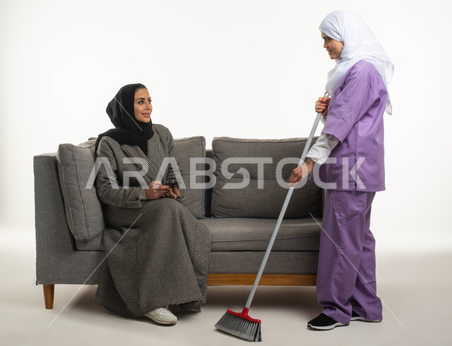 المهام والأعمال المنزلية الروتينية، أداء خدمة التنظيف تحت إشراف وتوجيه مدبرة المنزل، بورتريه لسيدة عربية خليجية سعودية محجبة تجلس على الأريكة تمسك بيدها الهاتف المحمول و تستعين بخادمة أجنبية داخل المنزل، عاملة نظافة تنظف بالمكنسة اليدوية، خلفية بيضاء