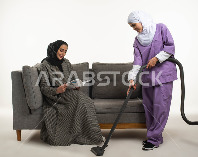 المهام والأعمال المنزلية الروتينية، أداء خدمة التنظيف تحت إشراف وتوجيه مدبرة المنزل، بورتريه لسيدة عربية خليجية سعودية محجبة تجلس على الأريكة وتقرأ الكتب تستعين بخادمة أجنبية داخل المنزل، عاملة نظافة تنظف بالمكنسة الكهربائية، خلفية بيضاء