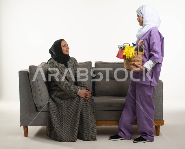 وظائف ومهن نسائية، بورتريه لعاملة نظافة أجنبية تحمل بيدها سلة قش بأدوات تنظيف المنزل تتلقى تعليمات النظافة من ربة المنزل، سيدة عربية خليجية سعودية محجبة تجلس على الأريكة وترشد الخادمة الإفريقية، خادمات منزلية مدربة ومؤهلة، خلفية بيضاء