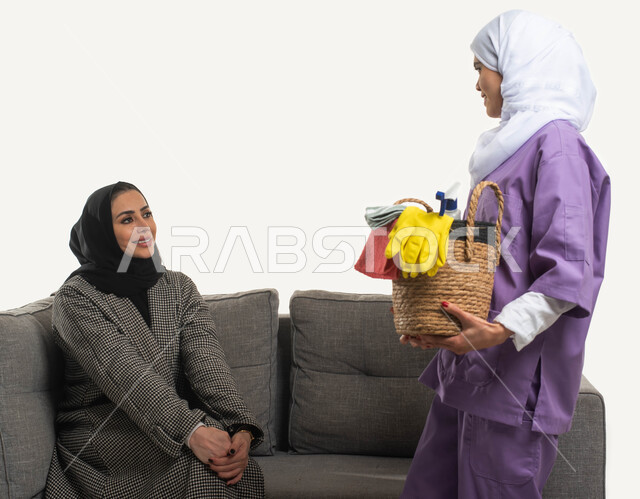 خادمات منزلية مدربة ومؤهلة، بورتريه مقرب لعاملة نظافة أجنبية تحمل بيدها سلة قش بأدوات تنظيف المنزل تتلقى تعليمات النظافة من ربة المنزل، سيدة عربية خليجية سعودية محجبة تجلس على الأريكة وترشد الخادمة الإفريقية، مهن ووظائف نسائية، خلفية بيضاء