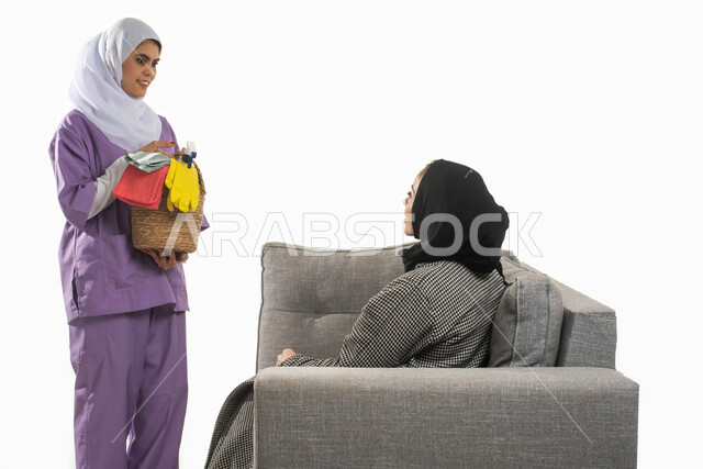 القيام بالمهام والأعمال المنزلية الروتينية، بورتريه لأمرأة عربية خليجية سعودية محجبة تجلس على الأريكة تستعين بخادمة أجنبية داخل المنزل، إعطاء التعلميات اللازمة لعاملة فلبينية تحمل بيدها سلة مليئة بمواد ومستلزمات التنظيف، خلفية بيضاء