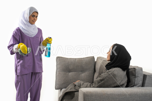 الحوارات والنقاشات مع عاملة فلبينية تحمل بيدها مواد ومستلزمات التعقيم والتنظيف، بورتريه من الجانب لأمرأة عربية خليجية سعودية محجبة تجلس على الأريكة تستعين بخادمة أجنبية داخل المنزل، أداء المهام والأعمال المنزلية الروتينية، خلفية بيضاء