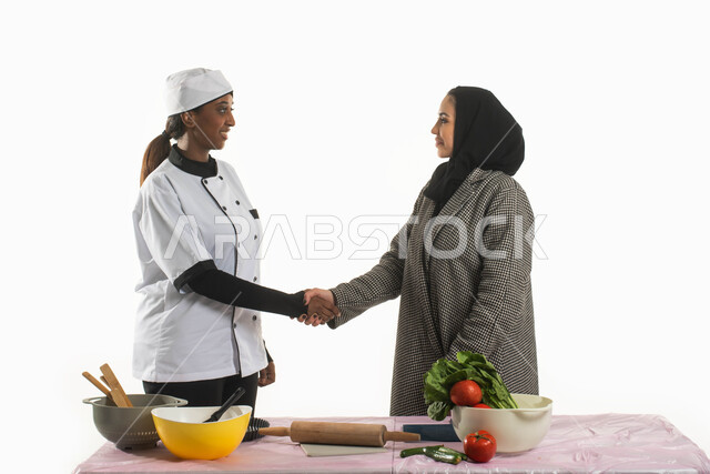 مجموعة من الخضار الطازجة على الطاولة والأدوات الخاصة بالمطبخ وأداة فرد العجين لتحضير الطعام، بورتريه لسيدة عربية خليجية سعودية محجبة تقوم بمصافحة طباخة فلبينية ترتدي مريول الطبخ، مفهوم إتباع النظام الغذائي الصحي والتدبير المنزلي، خلفية بيضاء