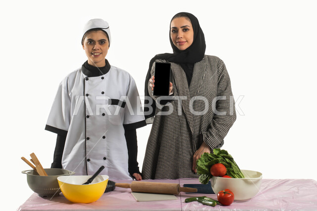 تسويق إعلانات للعمالة المنزلية، بورتريه لسيدة عربية خليجية سعودية محجبة تحمل بيدها الهاتف المحمول بجانبها شيف ترتدي مريول الطبخ، مجموعة من الخضار الطازجة على الطاولة والأدوات الخاصة بالمطبخ وأداة فرد العجين لتحضير الطعام، خلفية بيضاء
