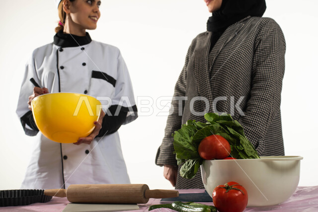 إعداد السلطات والأطباق الصحية، تدريب العاملات المنزلية على طريقة الطبخ العربي الخليجي، بورتريه مقرب لامرأة عربية خليجية سعودية محجبة تقوم بتعليم الطباخة المنزلية فنون المطبخ السعودي، وصفات وأكلات شعبية تقليدية، مهن ووظائف نسائية، خلفية بيضاء. 