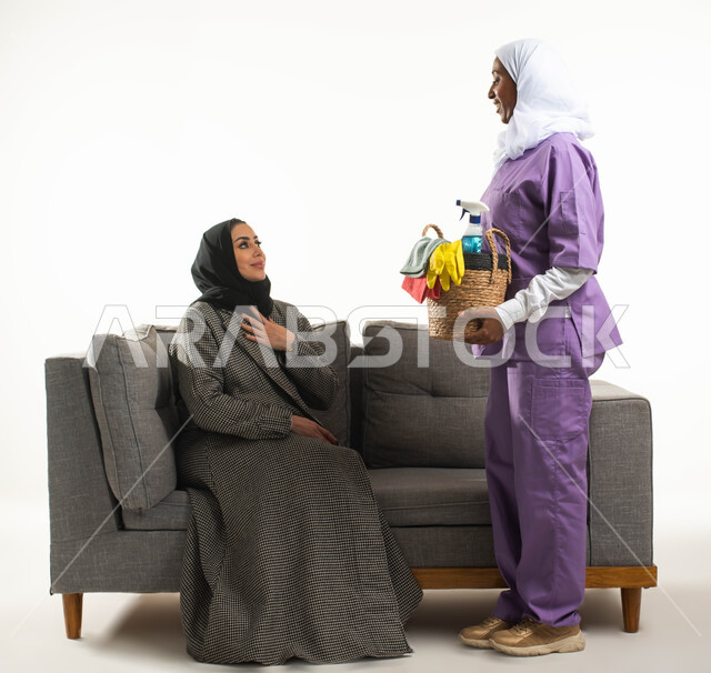 خادمات منزلية مدربة ومؤهلة، بورتريه لعاملة نظافة أجنبية تحمل بيدها سلة قش بأدوات تنظيف المنزل تتلقى تعليمات النظافة من ربة المنزل، سيدة عربية خليجية سعودية محجبة تجلس على الأريكة وترشد الخادمة الإفريقية، مهن ووظائف نسائية، خلفية بيضاء.