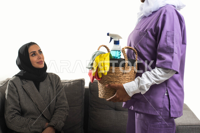 الاهتمام بتنظيف وتطهير البيت والقيام بالمهام الروتينية، عاملة نظافة أجنبية تحمل بيدها سلة قش بأدوات تنظيف المنزل، بورتريه مقرب لربة منزل عربية خليجية سعودية محجبة تجلس على الأريكة تقدم إرشادات النظافة للخادمة الإفريقية، مهن ووظائف نسائية، خلفية بيضاء.