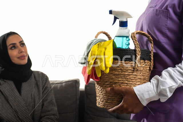 عاملة نظافة أجنبية تحمل بيدها سلة قش بأدوات تنظيف المنزل، التنظيف والتطهير والاهتمام بالأعمال المنزلية، بورتريه مقرب لربة منزل عربية خليجية سعودية محجبة تجلس على الأريكة تقدم إرشادات النظافة للخادمة الإفريقية، مهن ووظائف نسائية، خلفية بيضاء.