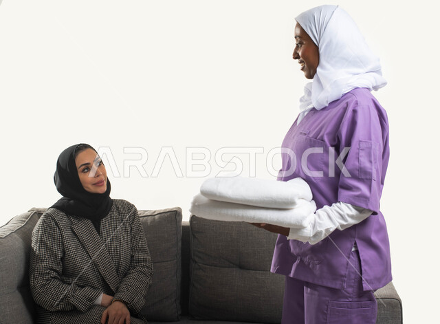 الإحسان في التعامل مع الخادمات في البيوت السعودية، طي وتنظيم الملابس وترتيبها، الاستعانة بعاملة أجنبية للقيام بالمهام المنزلية الروتينية، بورتريه مقرب لخادمة فلبينية تحمل بيدها منشفة نظيفة تقوم بمساعدة ربة منزل عربية خليجية سعودية محجبة، خلفية بيضاء. 