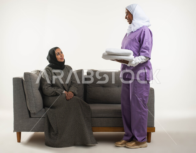تنظيف وطي وتنظيم الملابس وترتيبها، الاستعانة بعاملة أجنبية للقيام بالمهام والأعمال المنزلية الروتينية، بورتريه لربة منزل عربية خليجية سعودية محجبة تجلس على الأريكة وتعطي التعليمات اللازمة لخادمة إفريقية تحمل بيدها منشفة نظيفة، خلفية بيضاء. 