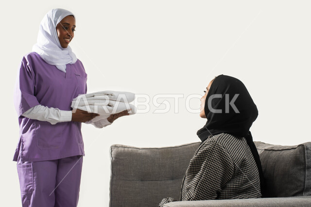 الاستعانة بعاملة أجنبية للقيام بالمهام والأعمال المنزلية الروتينية، بورتريه مقرب من الجانب لربة منزل عربية خليجية سعودية محجبة تعطي التعليمات اللازمة لخادمة إفريقية تحمل بيدها منشفة نظيفة، تنظيف وطي وتنظيم الملابس وترتيبها، خلفية بيضاء. 