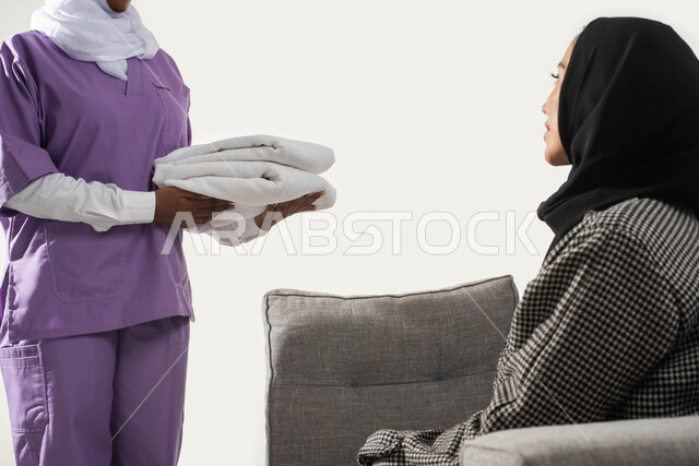 تنظيف وطي وتنظيم الملابس وترتيبها، الاستعانة بعاملة أجنبية للقيام بالمهام والأعمال المنزلية الروتينية، بورتريه مقرب من الجانب لربة منزل عربية خليجية سعودية محجبة تعطي التعليمات اللازمة لخادمة إفريقية تحمل بيدها منشفة نظيفة، خلفية بيضاء. 