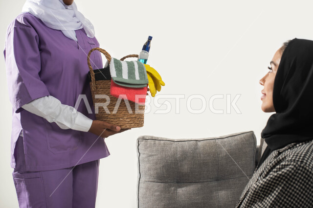 الاهتمام بالتنظيف والقيام بالمهام الروتينية، عاملة نظافة أجنبية تحمل بيدها سلة قش بأدوات تنظيف المنزل، بورتريه مقرب من الجانب لربة منزل عربية خليجية سعودية محجبة تجلس على الأريكة تقدم تعليمات النظافة للخادمة الإفريقية، مهن ووظائف نسائية، خلفية بيضاء.