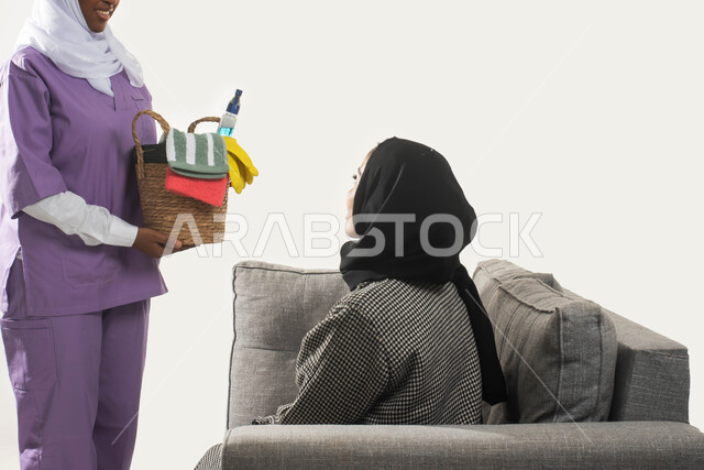 خادمات منزلية مدربة ومؤهلة، بورتريه مقرب من الجانب لعاملة نظافة أجنبية تحمل بيدها سلة قش بأدوات تنظيف المنزل تتلقى تعليمات النظافة من ربة المنزل، سيدة عربية خليجية سعودية محجبة تجلس على الأريكة وترشد الخادمة الإفريقية، مهن ووظائف نسائية، خلفية بيضاء.