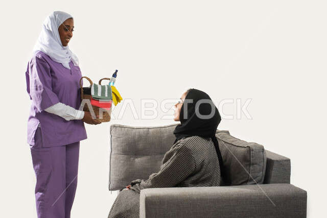 الخادمات في بيوت السعودية، بورتريه من الجانب لعاملة نظافة أجنبية تحمل بيدها سلة قش بأدوات تنظيف المنزل تتلقى تعليمات النظافة من ربة المنزل، سيدة عربية خليجية سعودية محجبة تجلس على الأريكة وترشد الخادمة الإفريقية، مهن ووظائف نسائية، خلفية بيضاء.