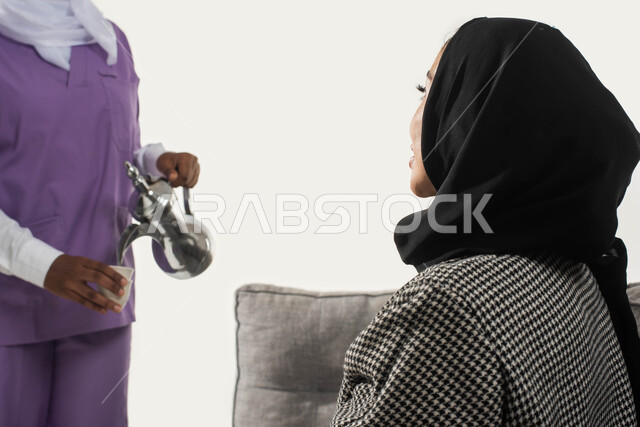 الخادمات في البيوت السعودية، القيام بالمهام والأعمال المنزلية الروتينية، حوارات ونقاشات مدبرة المنزل مع خادمة منزلية أجنبية، بورتريه مقرب من الجانب لسيدة عربية خليجية سعودية مع خادمة فلبينية تقوم بصب القهوة لتقديمها، حسن الضيافة والاستقبال، خلفية بيضاء.