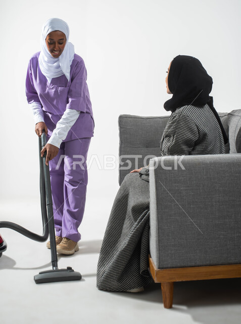 أداء خدمة التنظيف تحت إشراف وتوجيه مدبرة المنزل، بورتريه لسيدة عربية خليجية سعودية محجبة تستعين بخادمة أجنبية داخل المنزل، الحوارات والنقاشات، عاملة نظافة منزلية أجنبية تنظف بالمكنسة الكهربائية، الخادمات في البيوت السعودية، خلفية بيضاء.