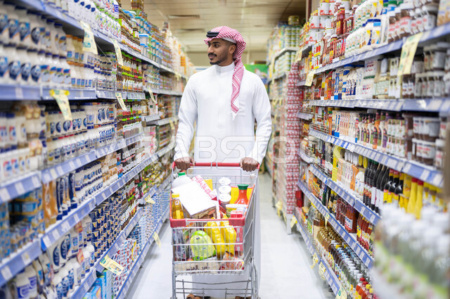 عرض منتجات محلية وطنية في الماركت, السهولة والراحة في التسوق, قضاء وقت ممتع في اختيار اللوازم المنزليه, امتلاك خيارات شراء و استهلاك مختلفة في السوبرماركت، رجل عربي سعودي خليجي يرتدي الثوب التقليدي و يمشي في أحد أقسام الماركت ويقوم بجر عربة التسوق