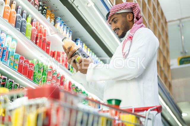 اختيار العصائر الطازجه المفيده، عرض المواد الغذائيه والتموينات المحليه والمستورده في الماركت, التسوق وشراء المستلزمات من متجر المواد الغذائية، رجل عربي سعودي خليجي يرتدي الثوب التقليدي يقوم بالتأكد من محتويات المنتج