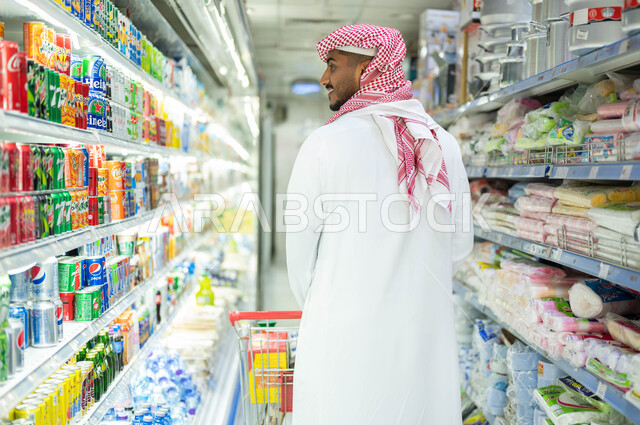 Possessing different purchase and consumption options in the mall, displaying local and national products in the market, ease and comfort in shopping, having a good time choosing household supplies, a back image of a Saudi Arabian Gulf man wearing the tra