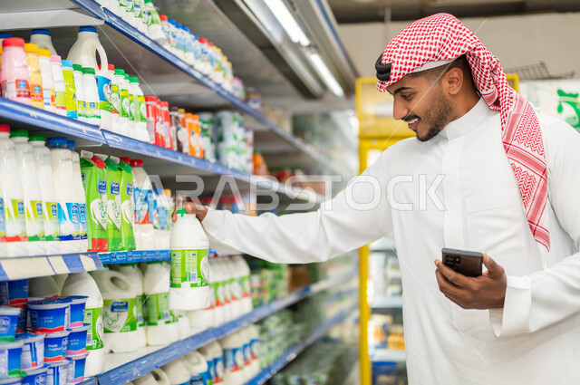 عرض منتجات وتموينات محلية ومستوردة في الماركت, شراء المتطلبات والاحتياجات الأساسية اليومية, اختيار بعض المستلزمات والمواد الغذائية الضرورية,  رجل عربي سعودي خليجي يبتسم يحمل بيده الهاتف المحمول يقوم بقراءة التعليمات من على علبة المنتجات