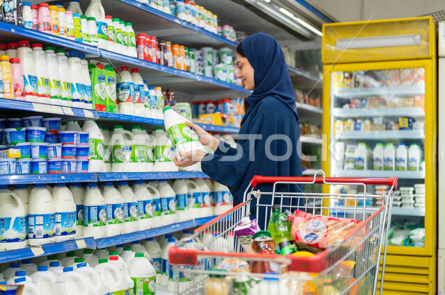 Purchasing daily basic requirements and needs, displaying local and imported products and supplies in the market, choosing some necessary supplies and needs, a side image of a smiling Saudi Arabian Gulf woman reading the instructions on the product box, t