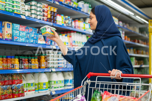 عرض منتجات وتموينات محلية ومستوردة في الماركت, اختيار وشراءعلبة من الجبن المحلي الطازج, إمرأة عربية خليجية سعودية مبتسمة تقوم بقراءة محتويات الاطعمة المعلبة وسعراتها الحرارية, قضاء وقت ممتع في شراء الاحتياجات الغذائية واقتناء المواد الغذائية