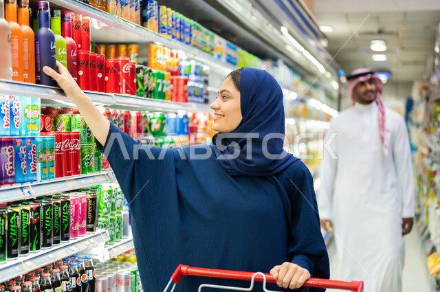  قضاء وقت ممتع في الماركت, تعابير وجه تدل على الاستمتاع بالتبضع والتسوق في المحلات التجارية,  سيدة عربية خليجية سعودية مبتسمة تقوم باختيار مشروب غازي, التسوق والحصول على المستلزمات المنزلية من متجر المواد الغذائية 
