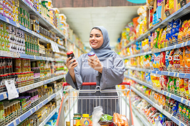 انتقاء الأفضل والأجود من السلع الموجودة في السوبر ماركت بالمملكة العربية السعودية، إمرأة عربية خليجية سعودية مبتسمة ترتدي العباءة الرمادية والحجاب ترتكز على عربة التسوق تمسك بيدها نوعين من المنتجات، ايماءات النظر إلى شيء ما، الاستفادة من العروض والتخفيضات