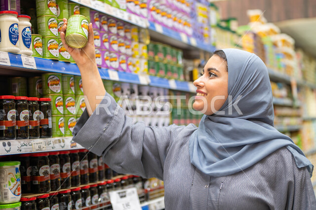 قضاء وقت ممتع في شراء المسلتزمات والمواد الغذائية، الاستفادة من التخفيضات والعروض والخصومات على المنتجات، سيدة عربية خليجية سعودية مبتسمة ترتدي العباءة الرمادية والحجاب تقوم باختيار وأخد المنتج، الاستعداد للتسوق وشراء الحاجات من المراكز التجارية السعوديه