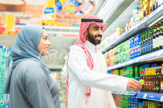 الاستمتاع بالتسوق وشراء المستلزمات، الاستفادة من التخفيضات والعروض والخصومات على المنتجات، زوجان عربيان خليجيان سعوديان مبتسمان يقومان باختيار المشروب الغازي، سيدة خليجية محجبة ترتدي العباءة الرمادية، رجل سعودي يرتدي الثوب والشماغ