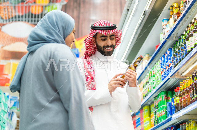 الاستفادة من التخفيضات والعروض والخصومات على المنتجات، زوجان عربيان خليجيان سعوديان مبتسمان يقومان بالتأكد من تاريخ صلاحية المنتج ومحتوياته، سيدة خليجية ترتدي العباءة الرمادية، رجل سعودي يرتدي الثوب والشماغ، الاستعداد للتسوق وشراء المستلزمات 