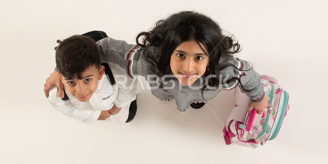 الاستعداد للعام الدراسي الجديد بايماءات تدل على السعادة، العلاقة الودية بين الأخوات, بورتريه علوي عامودي لأخوين عربيين سعوديين خليجيين يحملان الحقيبة المدرسية ينظران الى الكاميرا, مفهوم التعليم في السعودية والمثابرة على الدراسة لتحقيق النجاح, خلفية بيضاء