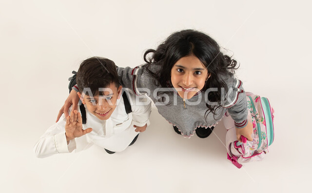 العلاقة الودية بين الأخوات, الاستعداد للعام الدراسي الجديد بايماءات تدل على السعادة، بورتريه علوي عامودي لأخوين عربيين سعوديين خليجيين يحملان الحقيبة المدرسية ينظران الى الكاميرا, مفهوم التعليم في السعودية والمثابرة على الدراسة لتحقيق النجاح, خلفية بيضاء