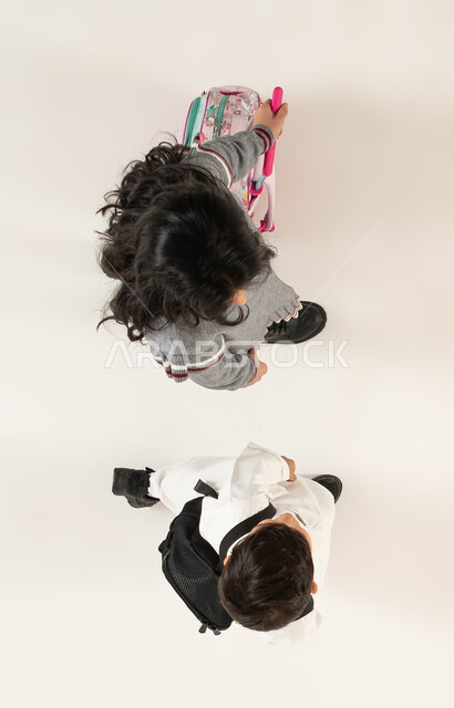 ايماءات السير والذهاب إلى المدرسة, الاستعداد للعام الدراسي الجديد، بورتريه علوي عامودي لولد وبنت عربيين سعوديين خليجيين يحملان حقيبة الظهر المدرسية, مفهوم التعليم في السعودية والمثابرة على الدراسة لتحقيق النجاح، خلفية بيضاء