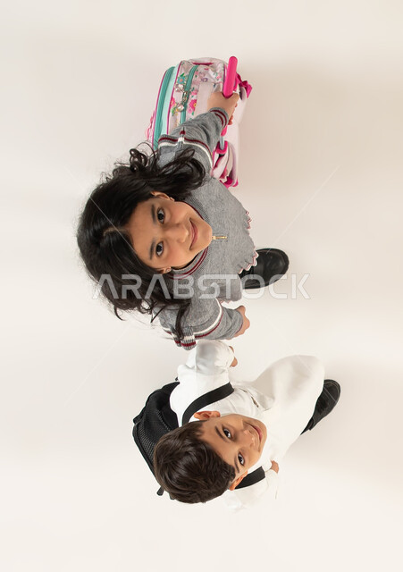 الاستعداد للعام الدراسي الجديد بايماءات تدل على الفرح والسعادة، بورتريه علوي عامودي لولد وبنت عربيين سعوديين خليجيين يحملان حقيبة الظهر المدرسية, مفهوم التعليم في السعودية والمثابرة على الدراسة لتحقيق النجاح، الذهاب إلى المدرسة, خلفية بيضاء