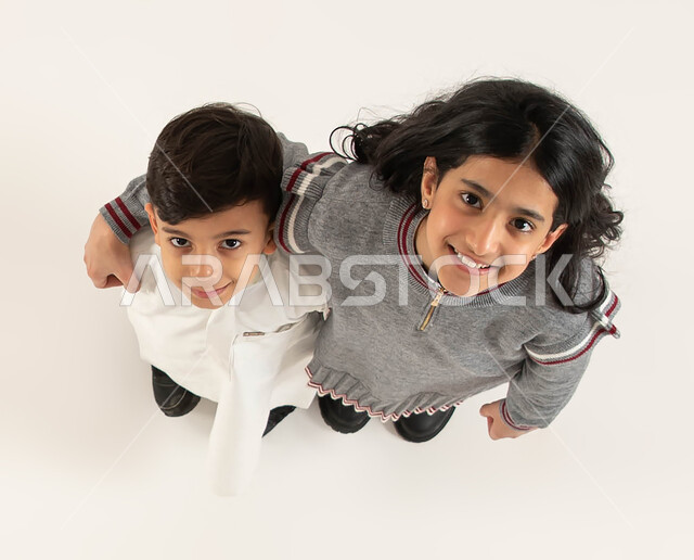 الاستمتاع بقضاء أوقات ممتعه برفقة الأخوات, تعزيز وتقوية العلاقة الإيجابية بين الأخوة, بورتريه علوي عامودي لأخويين عربيين سعوديين خليجيين  ينظران الى الكاميرا بايماءات السعادة والسرور, مفهوم الألفة والمحبة بين الأشقاء, خلفية بيضاء